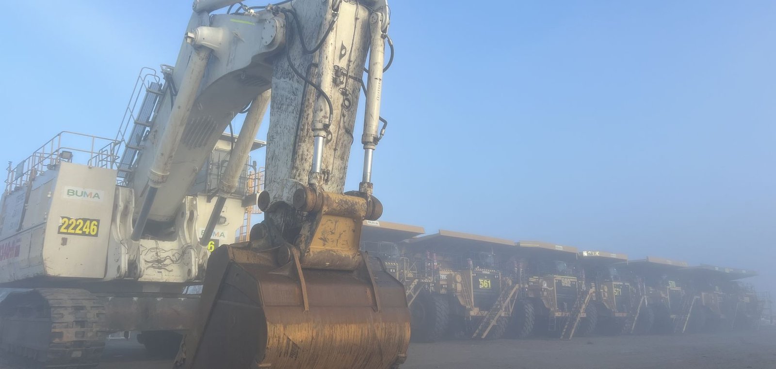 Mining excavator parked in heavy fog with a line of haul trucks behind it